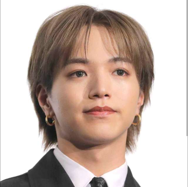 スーツ姿の男性が白背景の前で正面を向いているプロフィール写真。明るいブラウンの髪とゴールドのピアスが印象的。