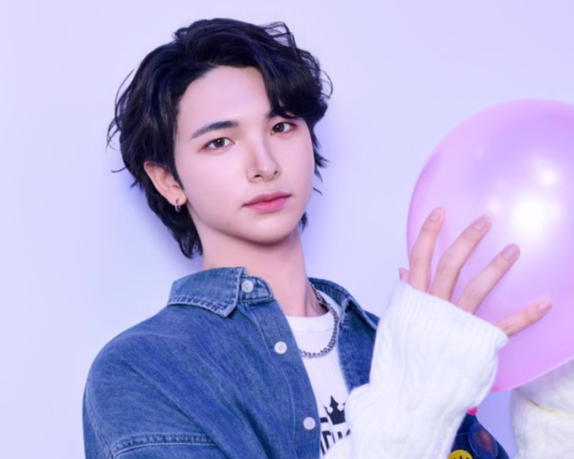 デニムシャツを羽織った黒髪の若い男性が、淡い紫色の背景でピンクの風船を手に持っている写真。