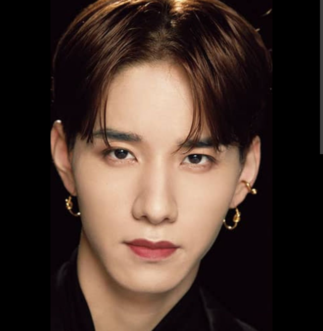 ダークカラーの衣装を着た人物の顔写真。センターパートの髪型で、両耳にピアスをつけ、黒い背景の前で正面を見つめている。