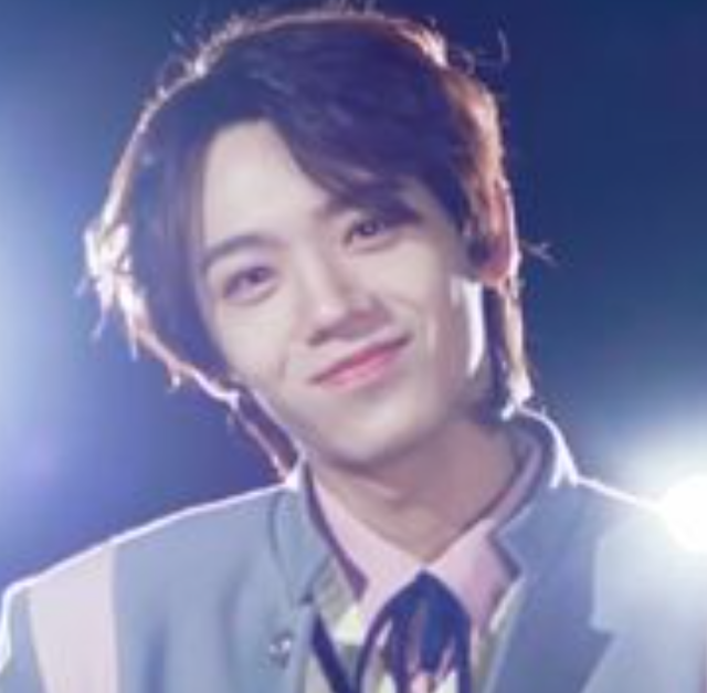 茶色がかった髪の人物が、ライトに照らされた中でやわらかく微笑んでいる上半身の写真。淡い色の衣装とリボンタイが印象