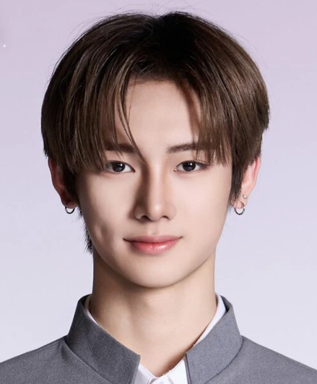 ブラウン系の髪をセンターパートにした人物が、グレーの制服風衣装を着て正面を向いているプロフィール写真。両耳のピアスとえくぼが印象的。