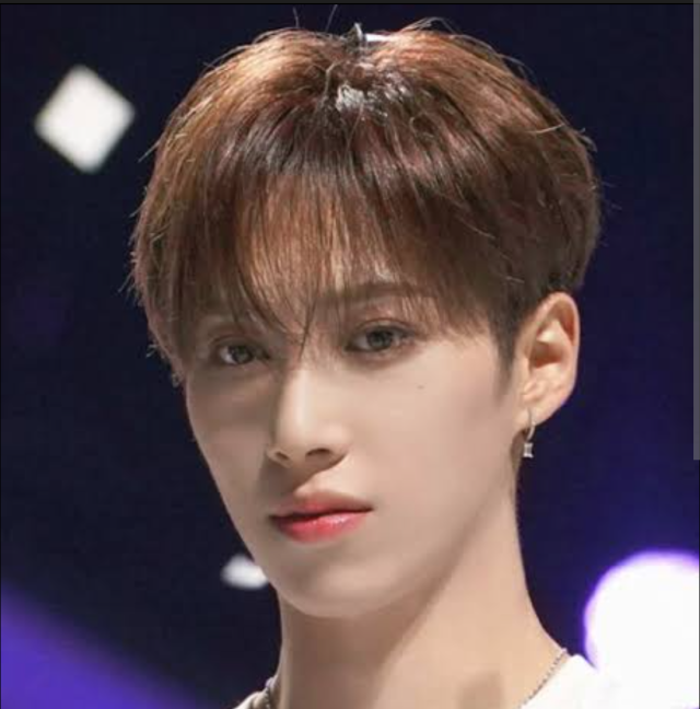 ブラウン系の髪をした人物が、ライトの入った暗めの背景の前で正面を見つめている写真。片耳のピアスと整った目元が印象的。