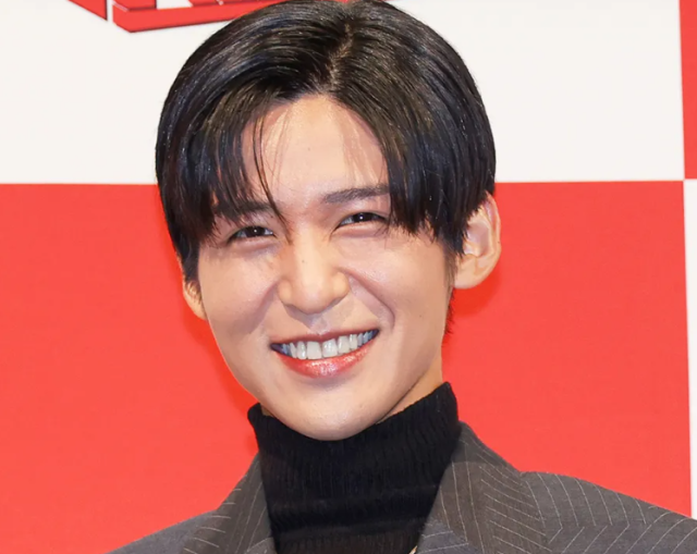 黒のタートルネックとジャケットを着た人物のポートレート写真。黒髪のセンターパートで、赤と白の背景の前で笑顔を見せている。