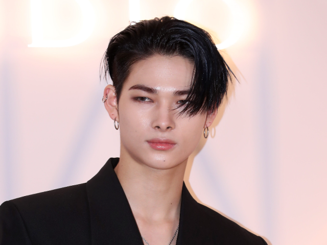 黒いジャケットを着た人物のポートレート写真。片目にかかる黒髪と複数のピアスが印象的で、明るい背景の前で正面を向いている。