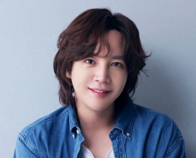 デニムシャツを羽織った人物のポートレート写真。やわらかな茶色がかった髪と穏やかな笑顔が印象的で、淡いグレーの背景の前に座っている。