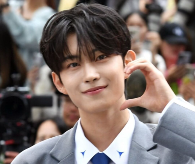 黒髪の若い男性がグレーのスーツ姿で微笑みながら、手でハートのポーズを作っている写真