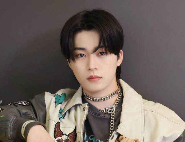 黒髪の若い男性がチェーンネックレスとジャケットを着け、落ち着いた表情で正面を見つめている写真