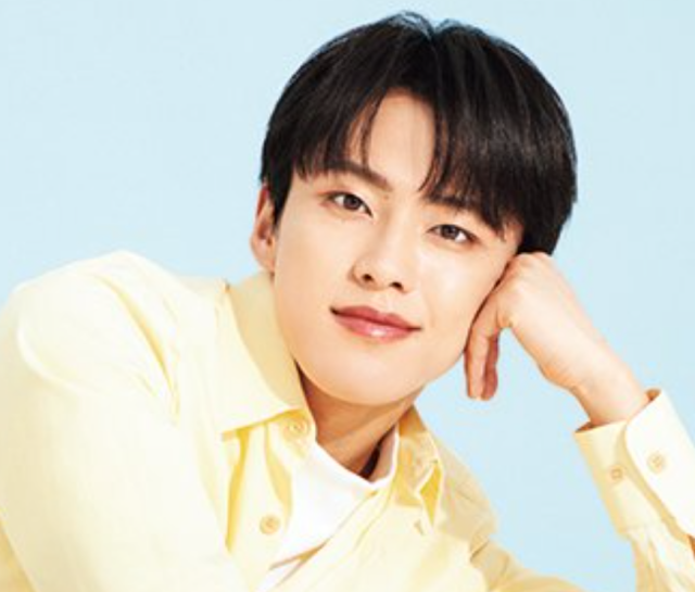 黒髪の若い男性が淡い黄色のシャツを着て、頬杖をつきながらやわらかく微笑んでいる写真