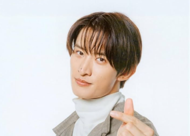 白背景の前で、グレーのジャケットと白いタートルネックを着た向井康二が、指ハートのポーズをしながらこちらを見ている画像。