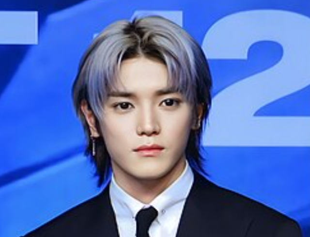 シルバーグレーの髪で黒いスーツとネクタイを着た人物が、青い背景の前で正面を向いている上半身画像