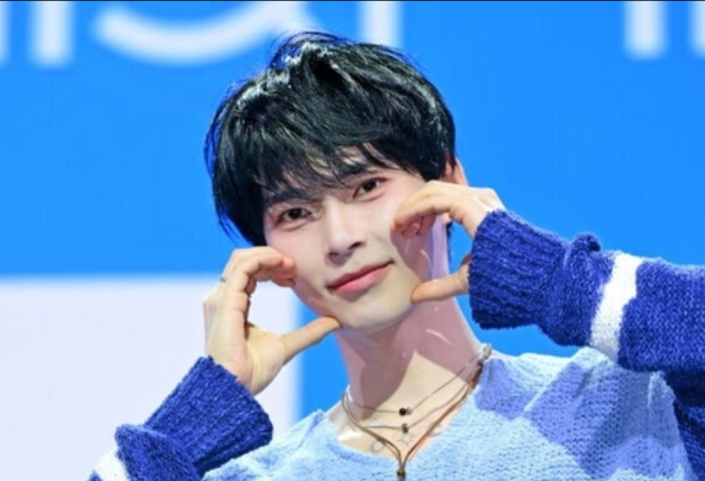 黒髪の人物が青いニット衣装を着て、両手で頬の横にハートポーズを作っているステージ写真